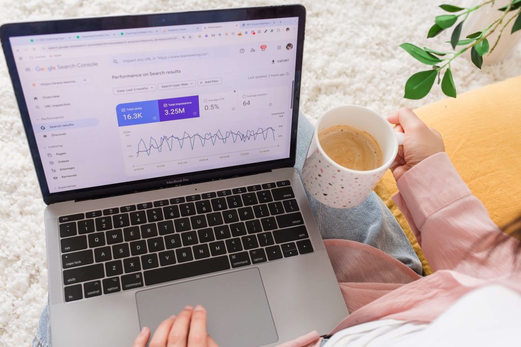 A woman holding a cup of coffee, typing away on her laptop. The laptop shows an impressive screenshot of Google Analytics