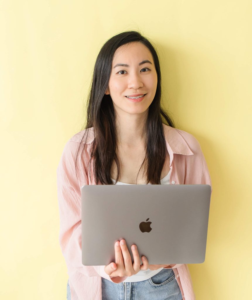 Founder Leanne Wong smiling in front of a yellow wall, holding her laptop